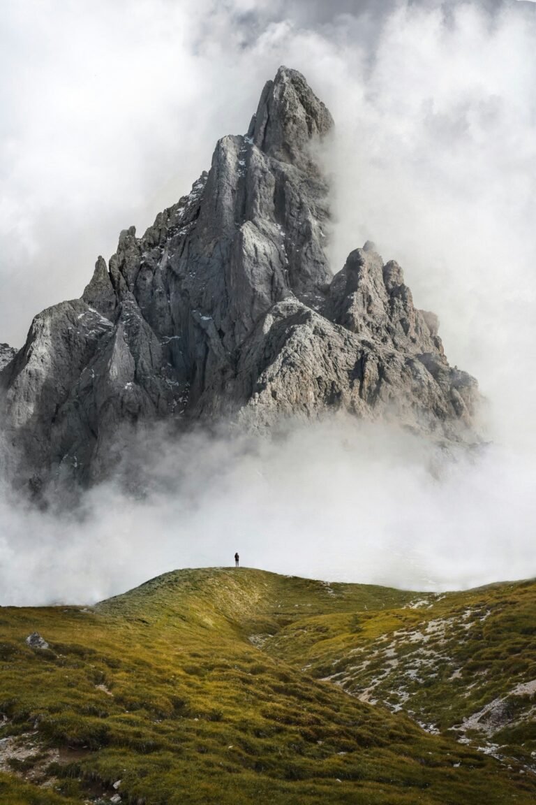 Top of a mountain surrounded by clouds, in the foreground there is a grassy hil and on top a person looking at the size of the mountain in front.